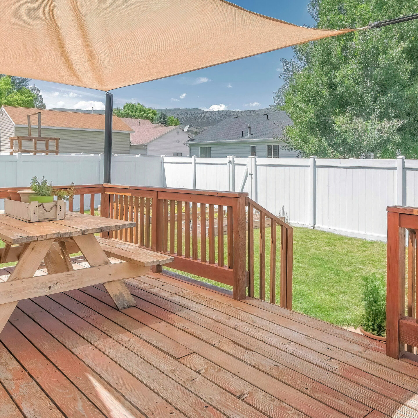 Panorama Whispy white clouds Wooden deck with a sunshade over the table with bench seats. Deck at the backyard of a house with a view of a lawn and white vinyl fence against the trees and neighborhood.
